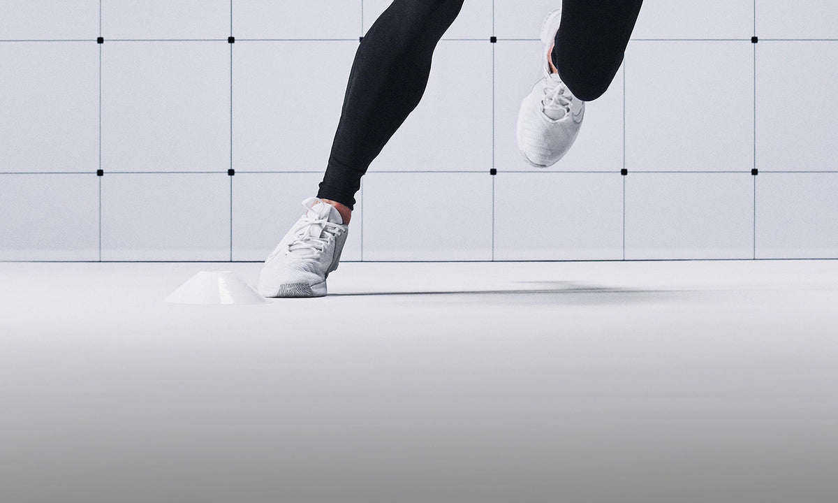 A person wearing black leggings and white athletic sneakers running against a minimalist white tiled wall and floor.