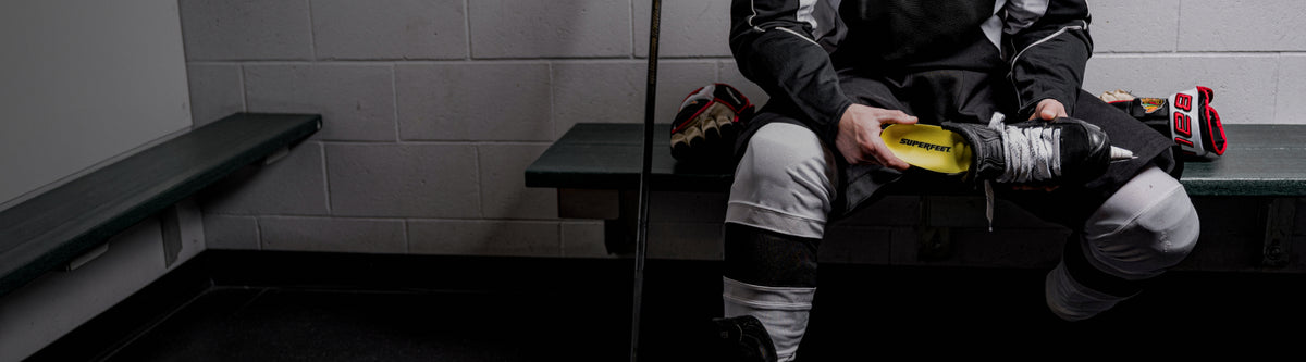 Person sitting on a bench in a hockey locker room with equipment. They are inserting a Superfeet Insole into their skates. 
