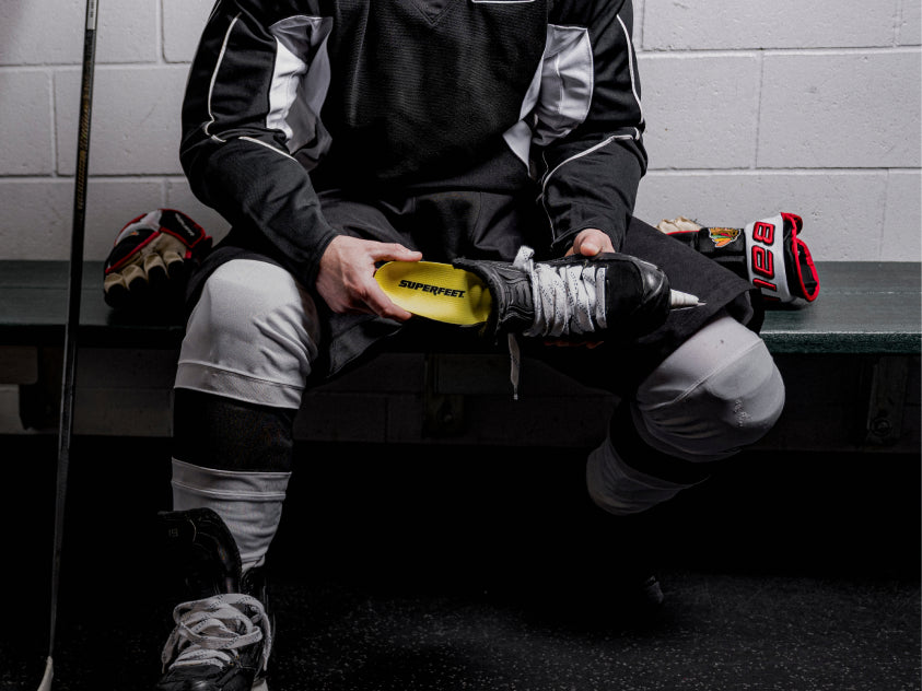Person sitting on a bench in a hockey locker room with equipment. They are inserting a Superfeet Insole into their skates. 