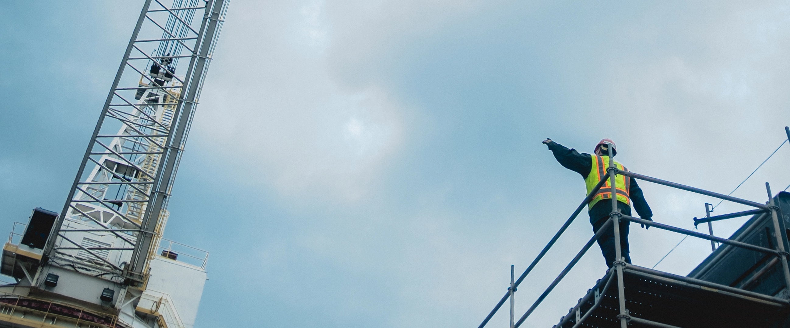 A person on scaffolding wearing a helmet & work safety vest while pointing out to a crane. 