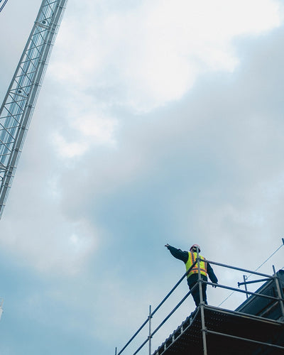 A person on scaffolding wearing a helmet & work safety vest while pointing out to a crane. 