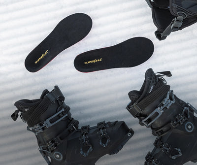 Black ski boots laid on some snow with Superfeet's Winter Think Support Insole laying near by. 