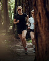 Two runners on a forest trail, one wearing sunglasses and a black outfit, the other in a white shirt and black shorts.