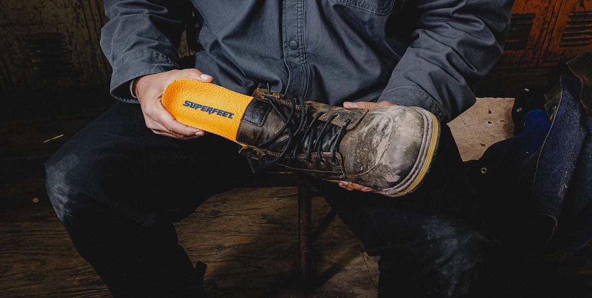 A person is sitting on a bench in an indoor setting, he is inserting Superfeet's Work Puncture Resistant Insole into a work boot. 
