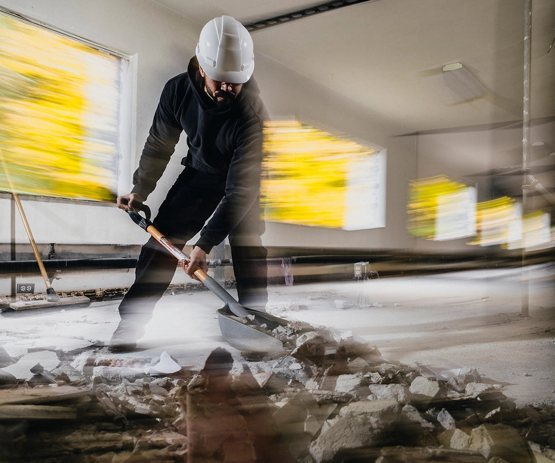 A blurry image of a man shoveling broken concrete with a shovel inside of an indoor construction site. 