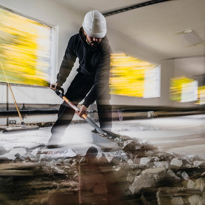 A blurry image of a man shoveling broken concrete with a shovel inside of an indoor construction site. 