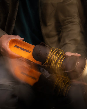 Someone inserting an orange 'Superfeet' insole into a a dark brown boot against a blurred background. 