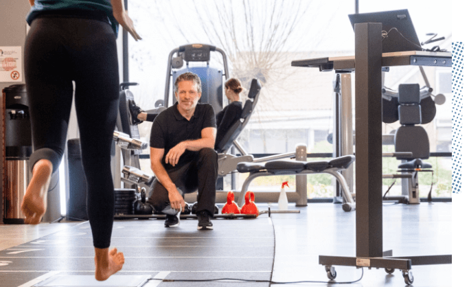 A practitioner in a well-equipped clinical gym setting attentively observes a patient sprinting across a gait scanner as the patient approaches.