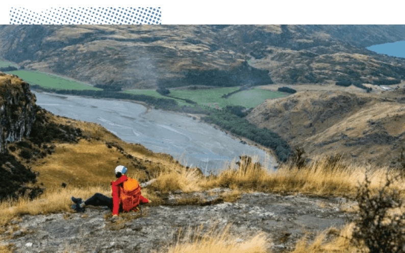 An adventurous person sitting on a mountainside, gazing at a picturesque valley with a wide winding river flowing through it.