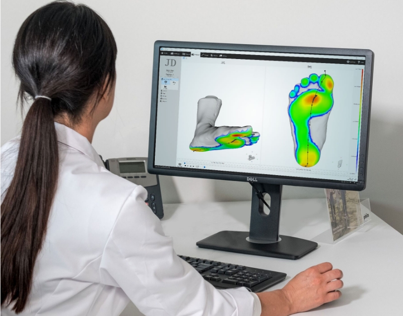 Practitioner seated and facing a computer screen with her back to the camera, examining a real-life scan of a patient's feet with visible heat maps representing pressure points. 