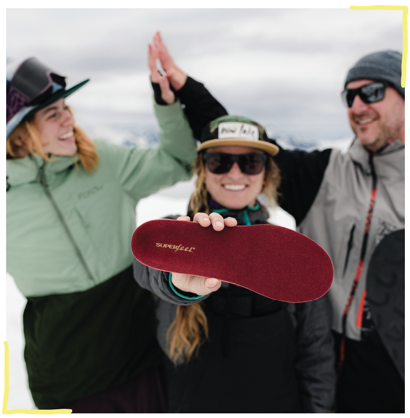 Three people in ski gear. Two people in background high five. Person in foreground holds Superfeet Winter Thin Support insole up to camera