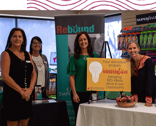 Four women standing behind a sign that says, "Your donation propels innovation bringing BIG ideas alive in our classrooms"